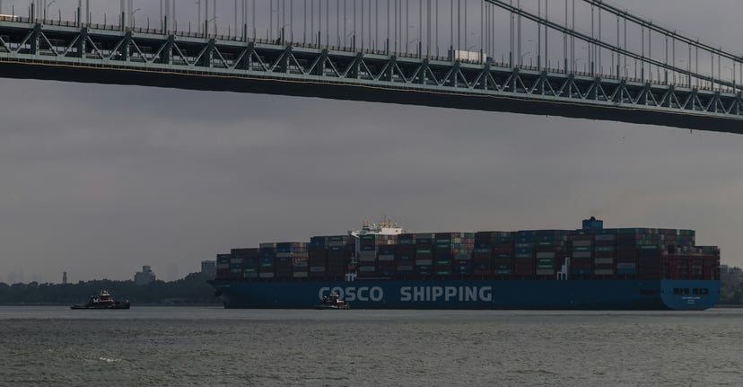 A COSCO Shipping container ship under grey skies and sailing past a suspension bridge