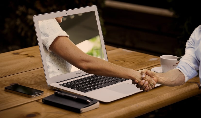 a hand reaching through a laptop screen to shake the hand of someone sitting at a desk