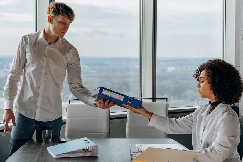 A young man in an untucked shirt handing an older employee a folder