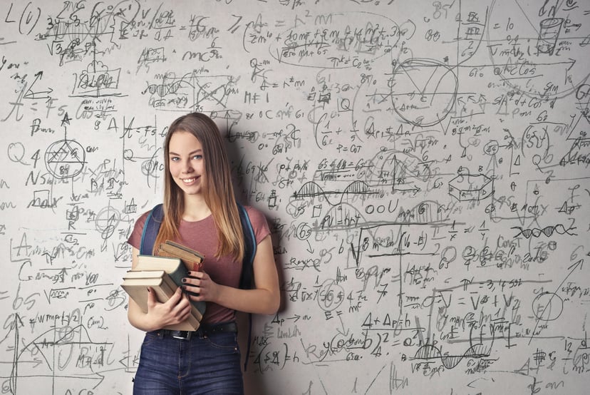 A young woman holding books and standing in front of a whiteboard covered in mathematical equations