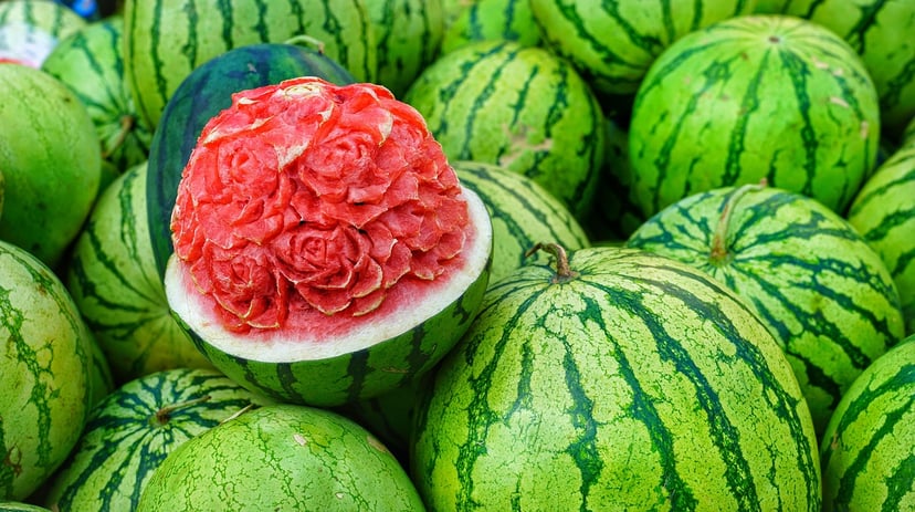 A pile of watermelons with one cut in half and carved into a bunch of roses