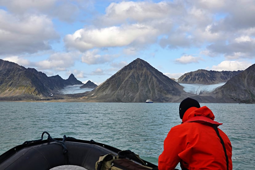 a man in a red jacket on the bow of a small dinghy heading towards small mountains