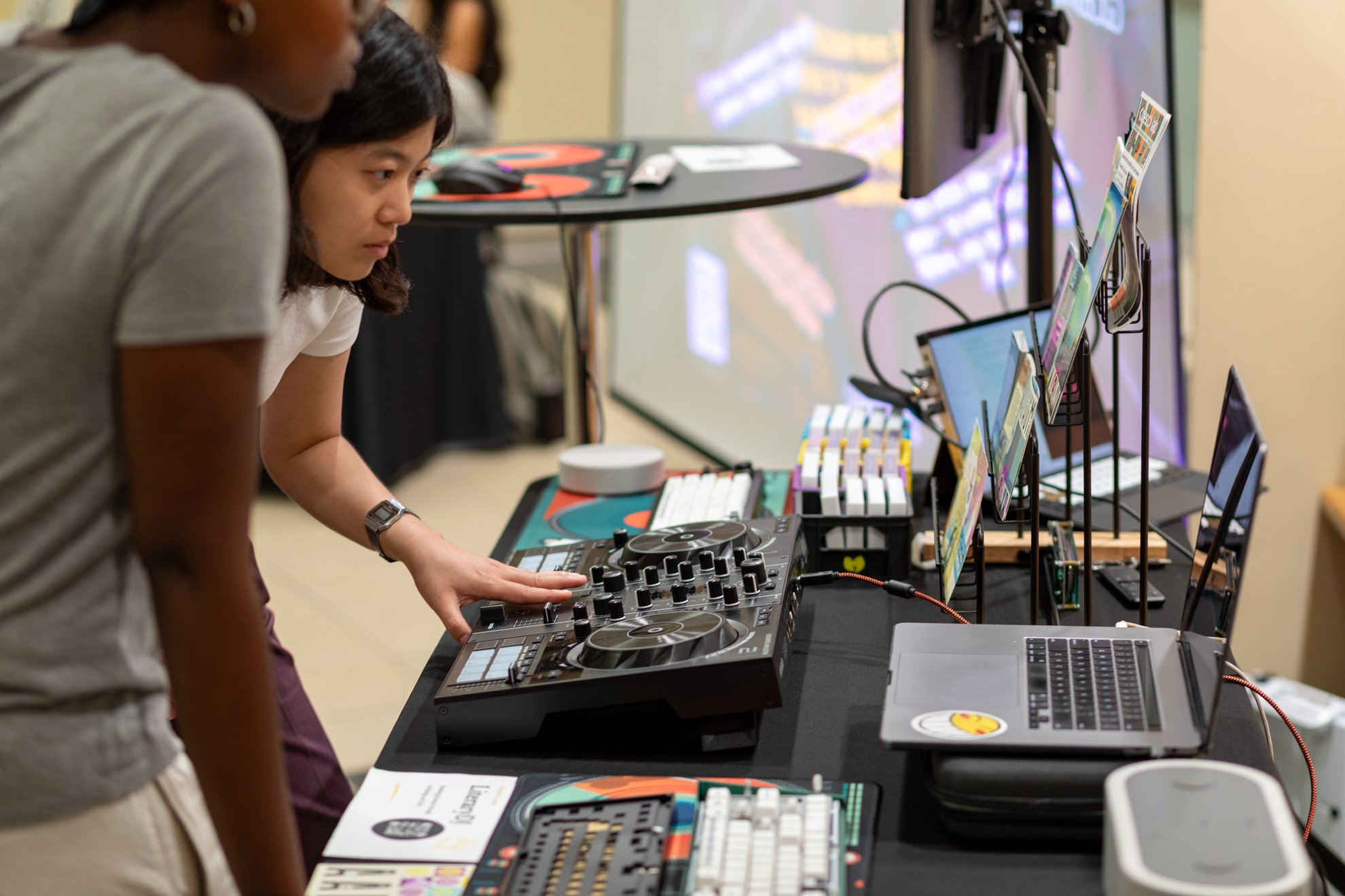 Two participants at a table of DJ controllers and keyboards, one with a hand on the controller.