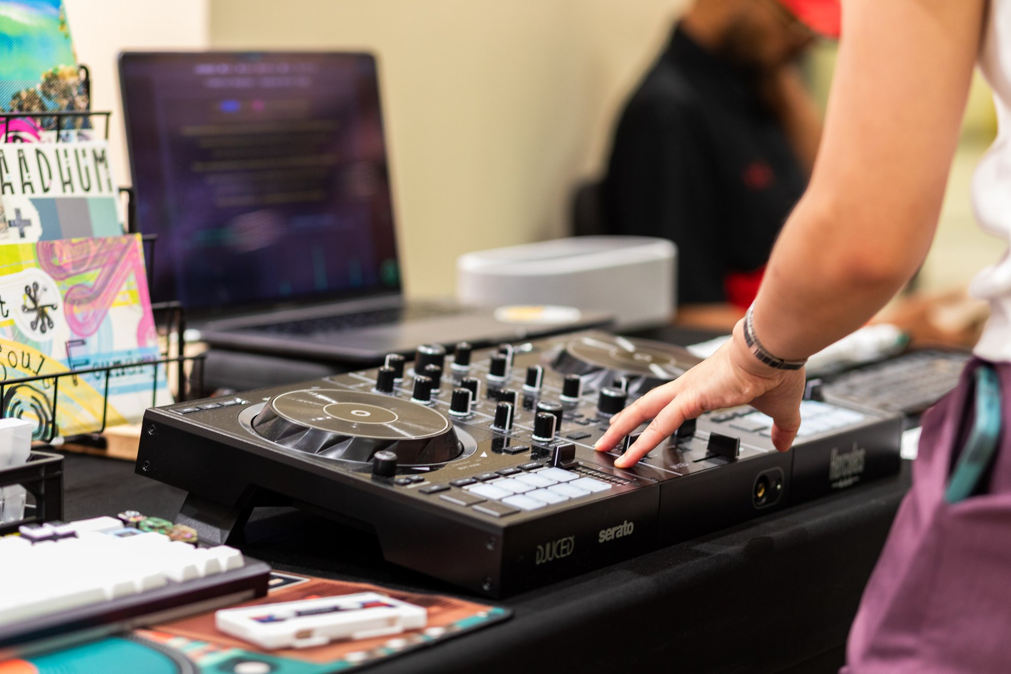 A hand pressing a pad on a Hercules DJ controller beside a laptop, with a custom keyboard and a cassette in the foreground.