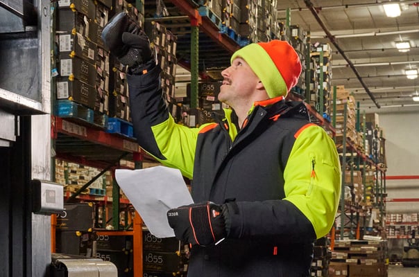 Man holds up a scan gun to scan a box in a warehouse filled with shelves stacked high that stretch out behind him. He's wearing  the FrostFlex Insulated Jacket combined wih the the FrostFlex Insulated Quilted Jacket to protect him from temperatures as low as -60°F. He's also wearing  PolarForce insulated work gloves and a reversible HiVis knit cap.