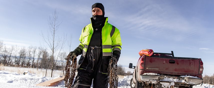 Man carrying snow chains wears a waterproof insulated jacket and bib overalls.