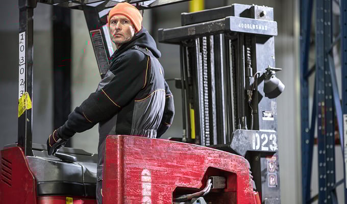 Man wearing an insulated sweatshirt and bib overalls drives a forklift in a refrigerated warehouse.