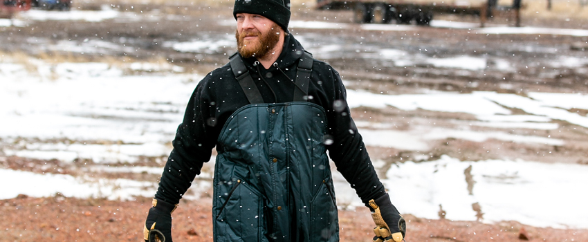 Man working outdoors in the winter wears insulated bib overalls, a thermal, hoodie and insulated work gloves.