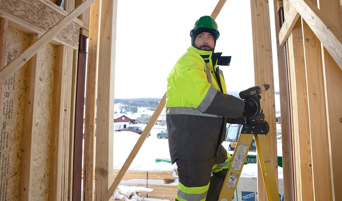 Man works on a snowy construction jobsite wearing an insulated jacket and bib overalls.