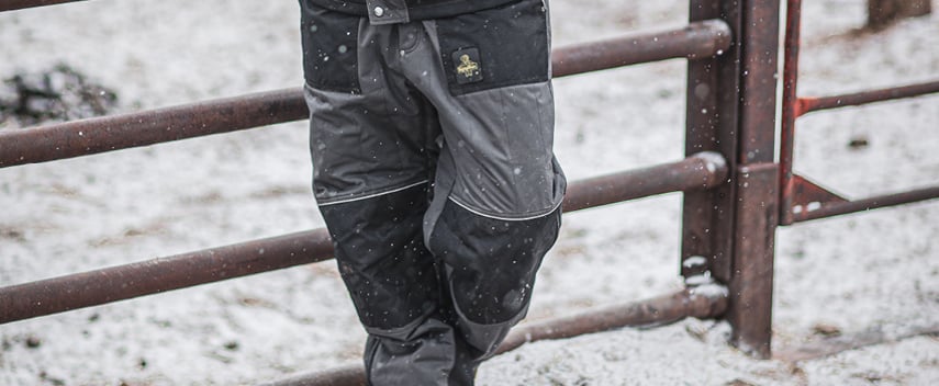 Person wearing gray and black work pants  stands by a fence in the snow.