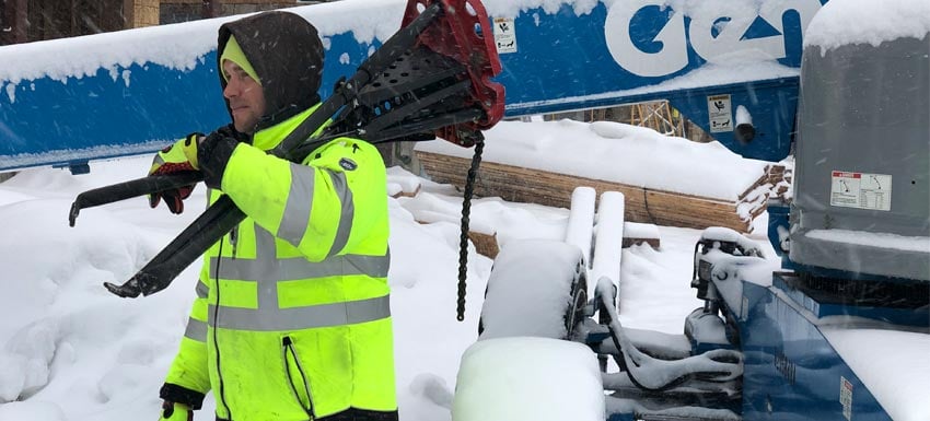 A man in a HiVis jacket carries long metal pieces over his shoulder in front of a large blue crane arm. Everything is covered in snow.