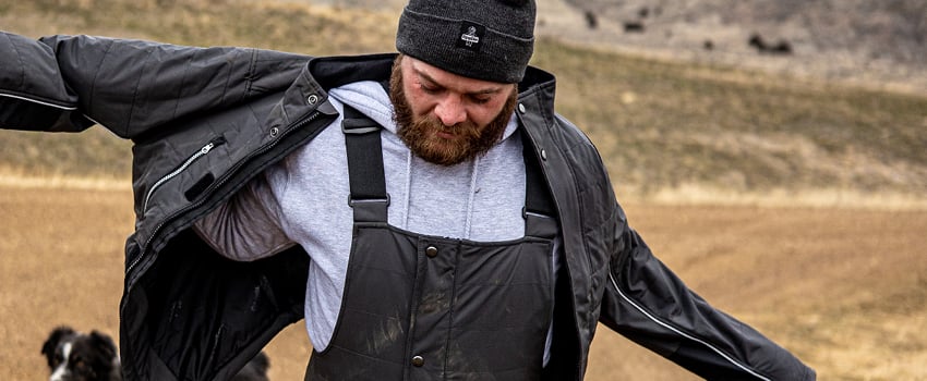 Man working outside in cold weather wears multiple layers, including a thermal base layer, a sweatshirt, insulated bib overalls and a winter work coat.