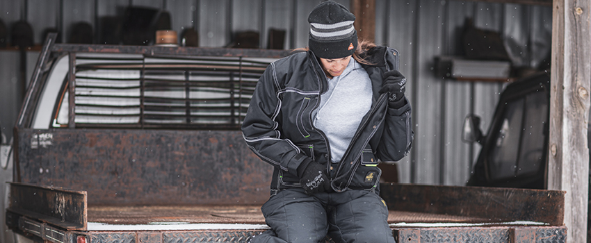 Woman working outside in a garage stays warm with multiple layers, including a sweatshirt, a waterproof work jacket, and insulated work pants.