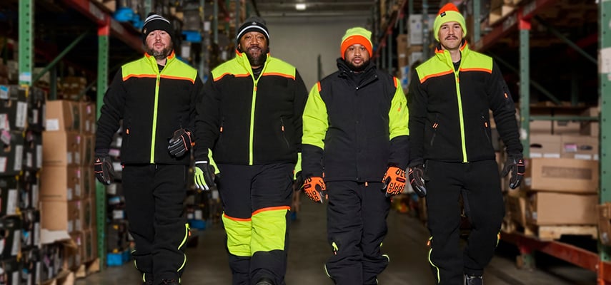 Four men walk toward the camera wearing FrostFlex insulated workwear from RefrigiWear in a cold storage warehouse. They're dressed for the freezer warehouse in FrostFlex insulated workwear from  RefrigiWear, including insulated work jackets and heavy-duty bib overalls, all with high visibility panels and piping that keep them safe and seen.