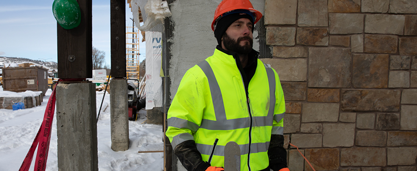 Man working on a snowy construction jobsite wears a thermal high-visibility jacket and a knit cap under a hard hat.