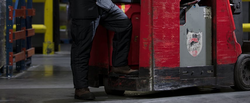 Person wearing insulated work pants steps onto a fork lift in a refrigerated warehouse.