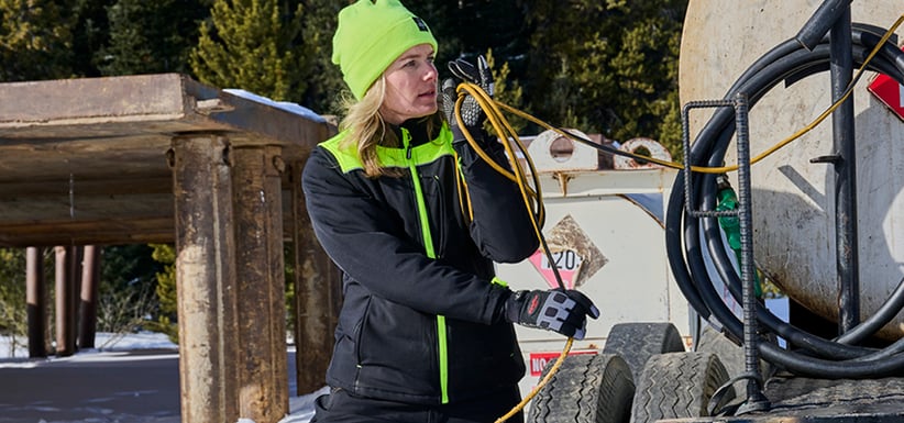 A blond woman wearing an insulated jacket with high-visibility lime shoulder patches, insulated work gloves and a HiVis knit cap wraps a cable around her arm on an outdoor job site.  Behind her is a high wooden platform, industrial equipment and beyond that, a pine forest.
