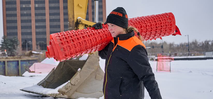 Hardworking woman in high-visibility women’s workwear hauling safety fencing on a snowy construction site—because female PPE isn’t just pink, it’s built tough.