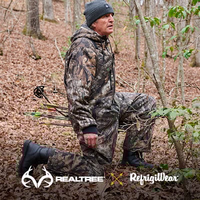 Man on an archery hunt kneels while wearing RefrigiWear Realtree APX Camo insulated overalls and sweatshirt.