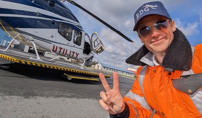 Smiling and clean-shaven, Mark’s gaze is shaded by a baseball cap as he looks into the camera through mirrored aviator sunglasses and takes a selfie, holding up two fingers on his right hand in a peace sign. Behind him we see a helicopter waiting with the door open, as though Mark’s about to jump in and take off. It’s a brisk winter day and Mark’s dressed in cold weather gear, wearing HiVis insulated coveralls from RefrigiWear with his collared shirt sticking out at the top. His left chest pocket has a black triangular flap that reads “IRON-TUFF” and closes with a brass snap. The sky is sunny and blue with fluffy white clouds and it looks like a great day to fly a helicopter.