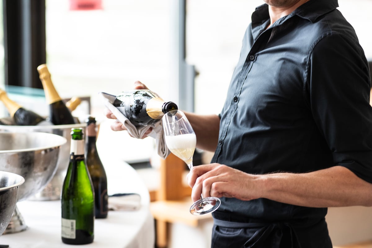 Sekt wird professionell eingeschenkt. Ein Glas Sekt wird in der Hand gehalten. Sektkühler im Hintergrund.