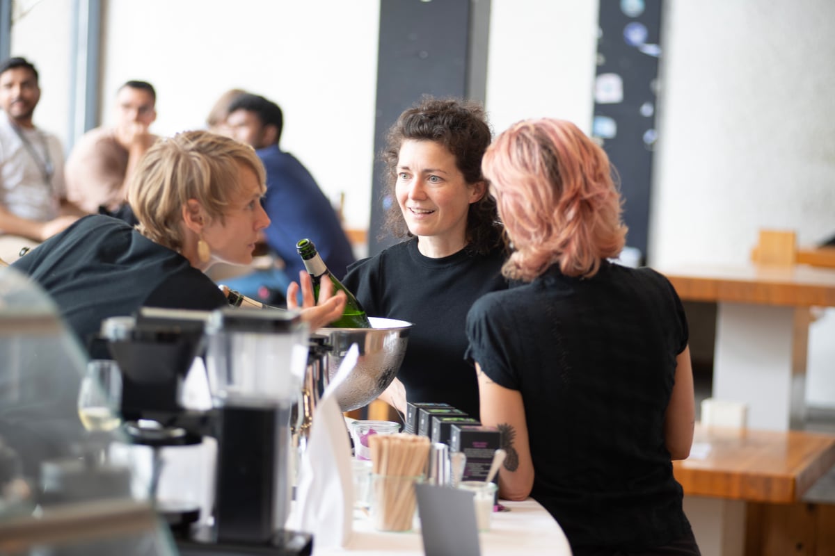Alexandra mit Servicekräften und Gästen im Hintergrund am Thresen. Sektkühler und Gläser sind bereit.