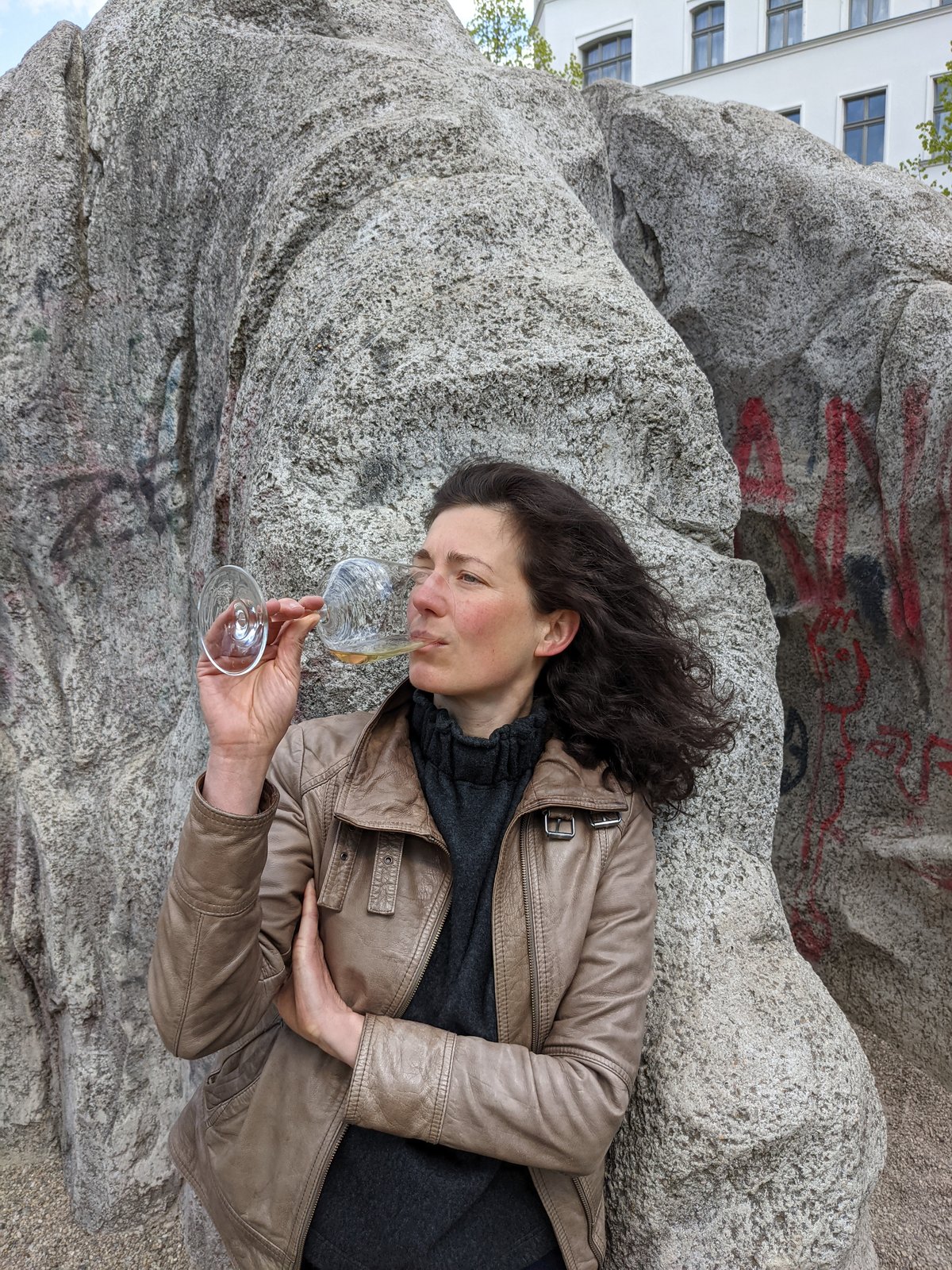 Alexandra in brauner Lederjacke vor Klettersteinen. Aus Glas trinkend.