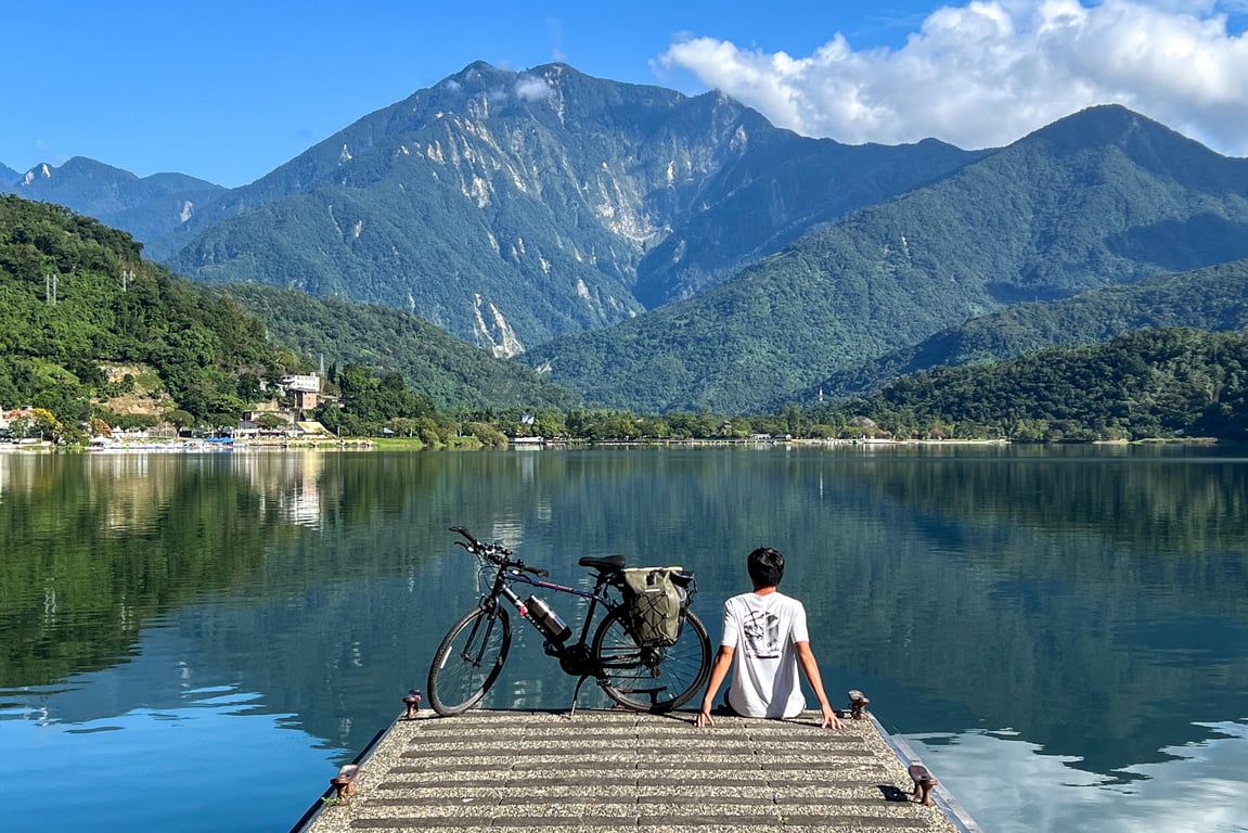 A cyclist sits on a wooden pier beside a touring bike, looking across the still water of Liyu Lake to Hualien's coastal mountains
