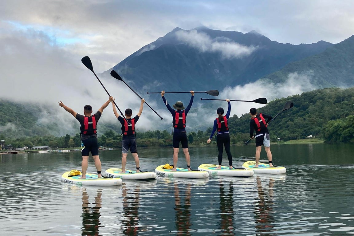 Five paddlers on stand-up paddleboards line up on the flat water of Liyu Lake, Hualien, paddles raised overhead with a cloud-wrapped ridge behind them