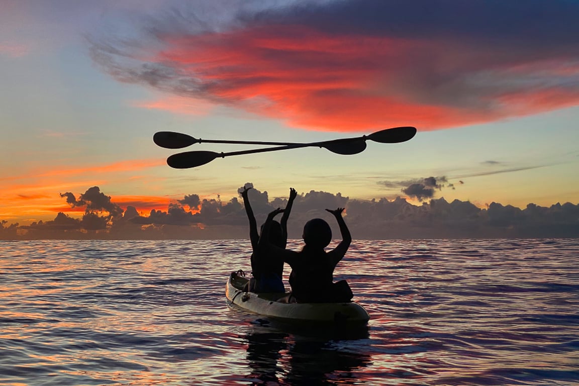 Two paddlers on a sea kayak raise their paddles overhead against a Pacific sunset, silhouetted below Qingshui Cliff, Hualien