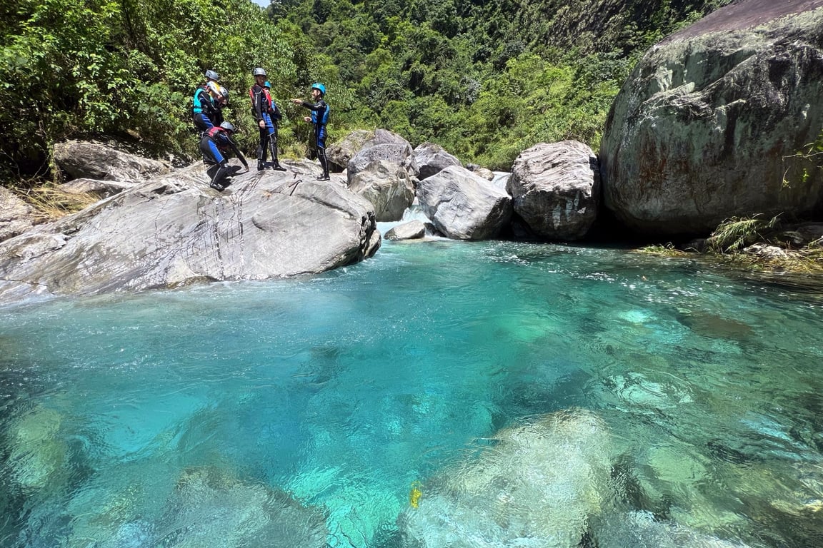 A group of river tracers in helmets and wetsuits stand on large marble boulders above a glowing turquoise pool — Baibao Creek, Hualien