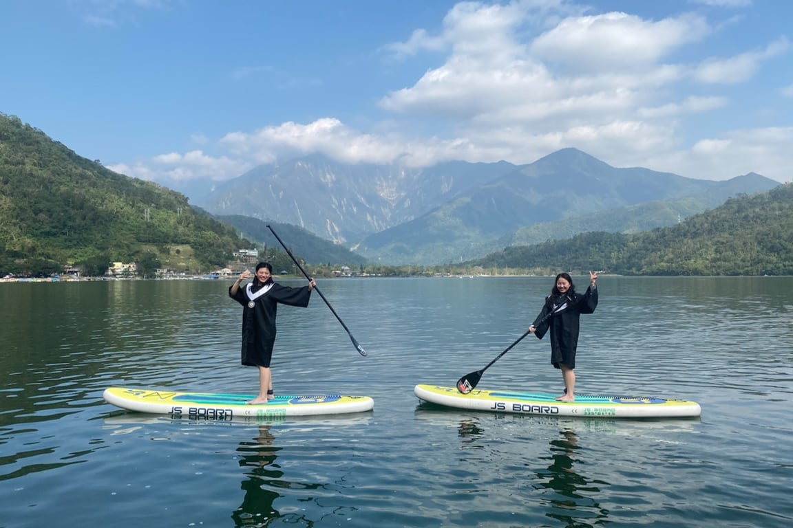 兩位女生穿著黑色學士服站在鯉魚潭 SUP 板上比 V, 背景是中央山脈藍天白雲