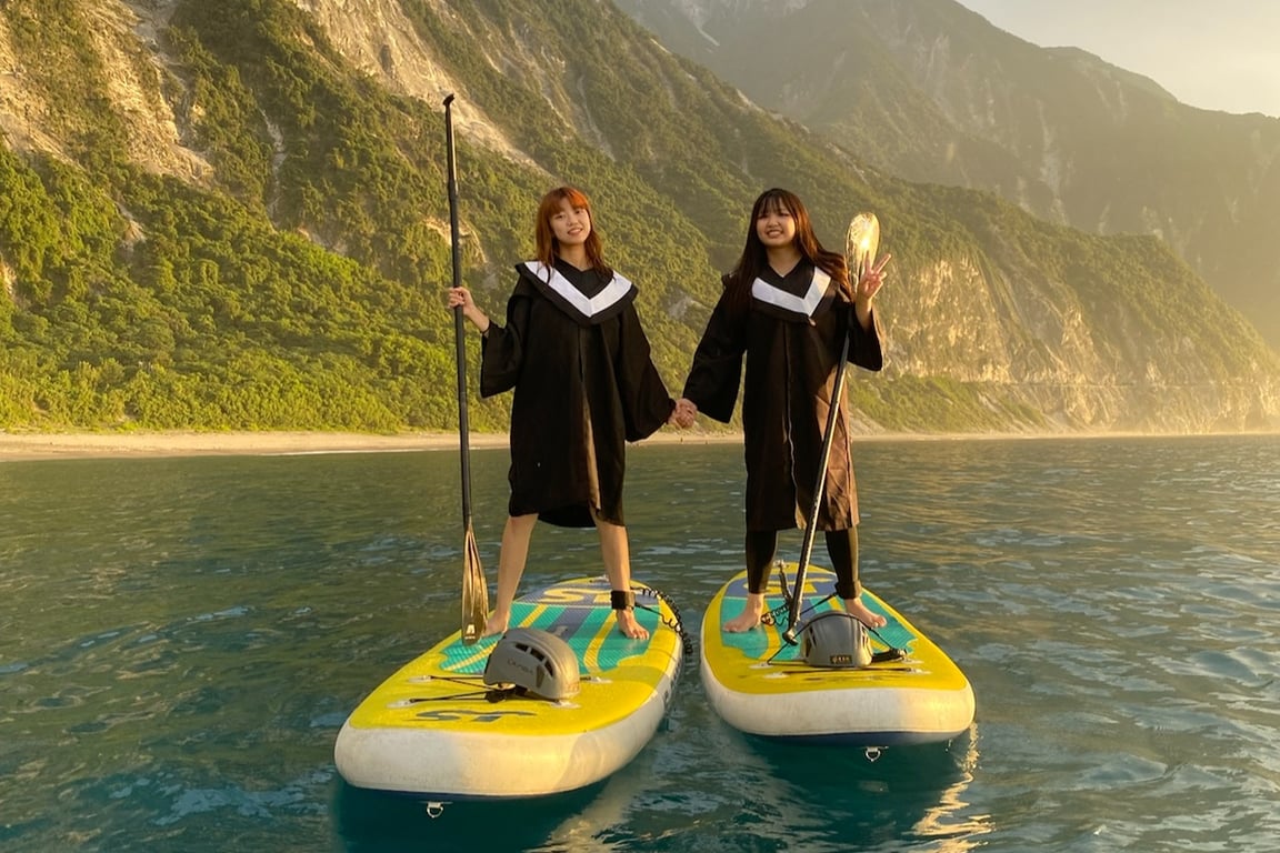 兩位女生穿著黑色學士服站在清水斷崖海面上的 SUP 板, 牽手 + 比 V, 背景是金色夕陽下的崖壁與綠林