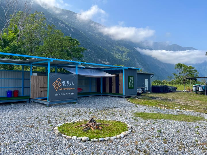 愛呆玩崇德基地外觀 ── 藍色屋簷的半戶外建築, 前方有石頭圍成的營火圈, 後方雲霧繚繞的山稜