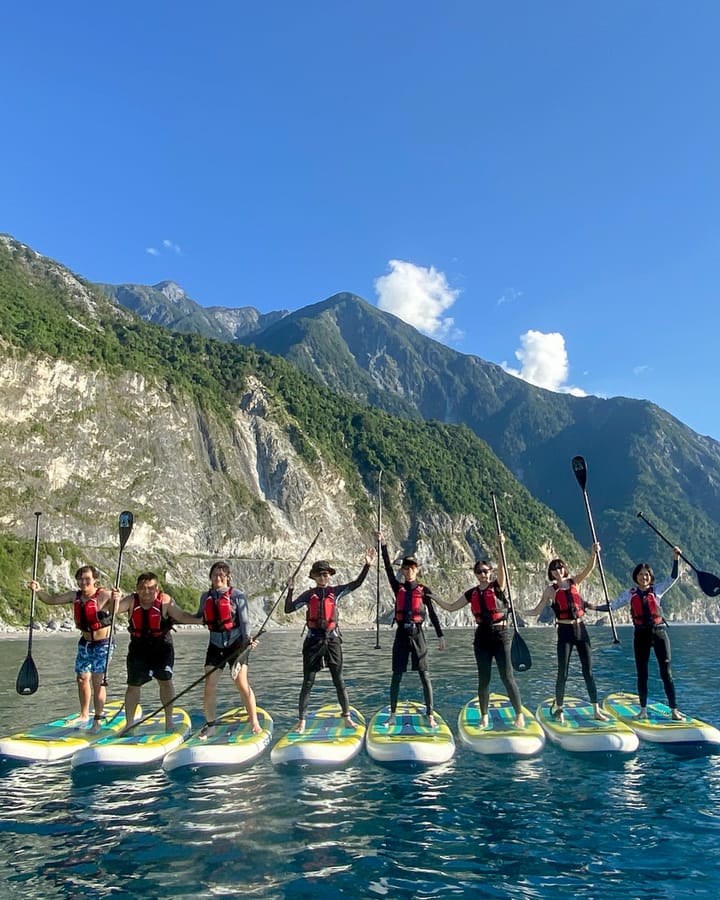 八位學員並排站在 SUP 板上比 V, 背景是清水斷崖陡峭岩壁與翠綠山稜, 太平洋藍天遼闊