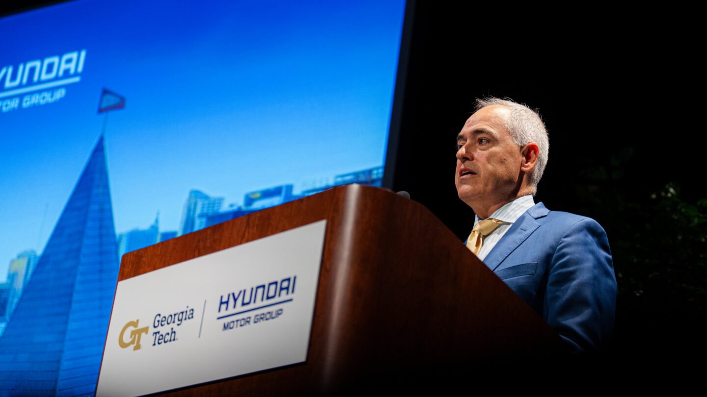 A man speaks behind a podium in front of a blue projected background