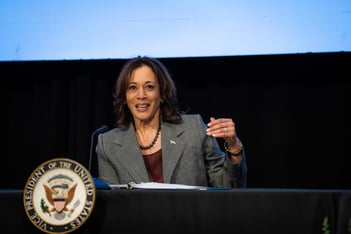 Vice President Kamala Harris delivers remarks while sitting at a table, leaning into a microphone. The seal of the vice president is in front of the table where she sits.