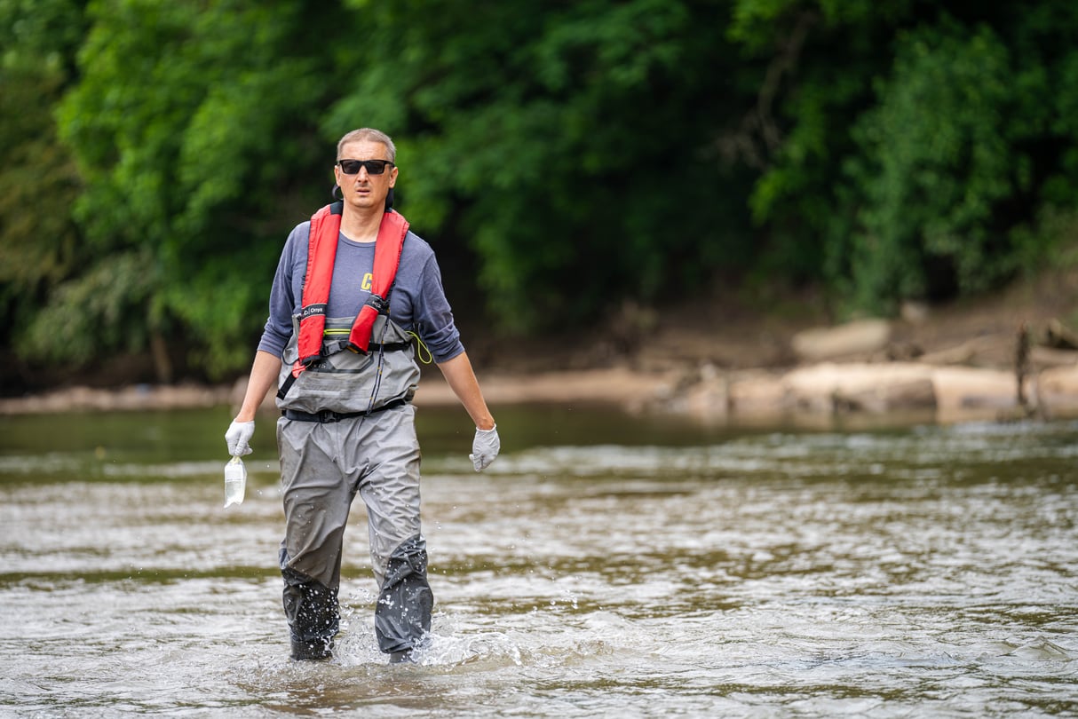 Jason Ulseth, wearing an orange PFD and grey waiters on top of a blue, long sleeve shirt wades through knee height water in the Chattahoochee.
