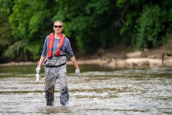 Jason Ulseth, wearing an orange PFD and grey waiters on top of a blue, long sleeve shirt wades through knee height water in the Chattahoochee.