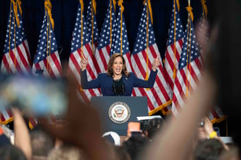 Vice Kamala Harris raises her hands at a rally at West Allis Central High School. A row of American flags stand behind her.