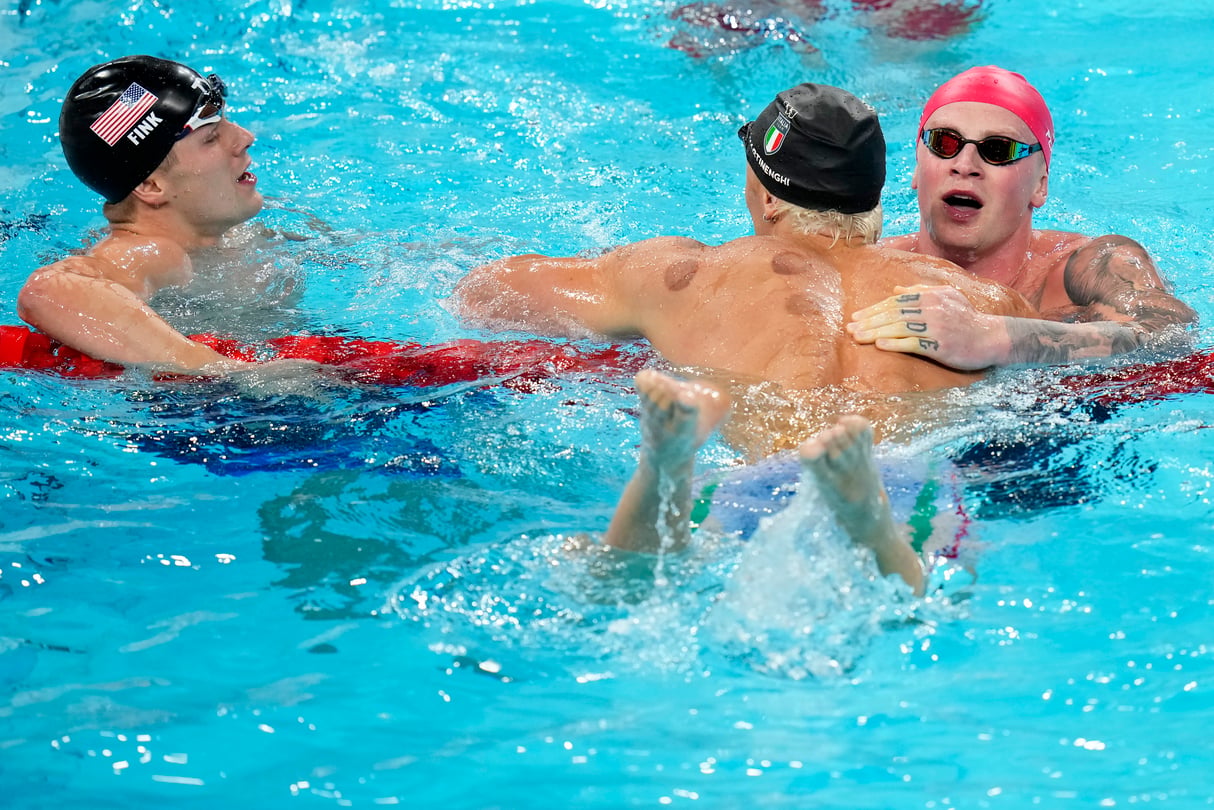 Nicolo Martinenghi, center, of Italy, is congratulated by Adam Peaty, of Great Britain, right, and Nic Fink, of the United States, left, after winning the men's 100-meter breaststroke final at the 2024 Summer Olympics on Sunday. All three men are hovered over the lane line in the pool, cap and goggles still on.