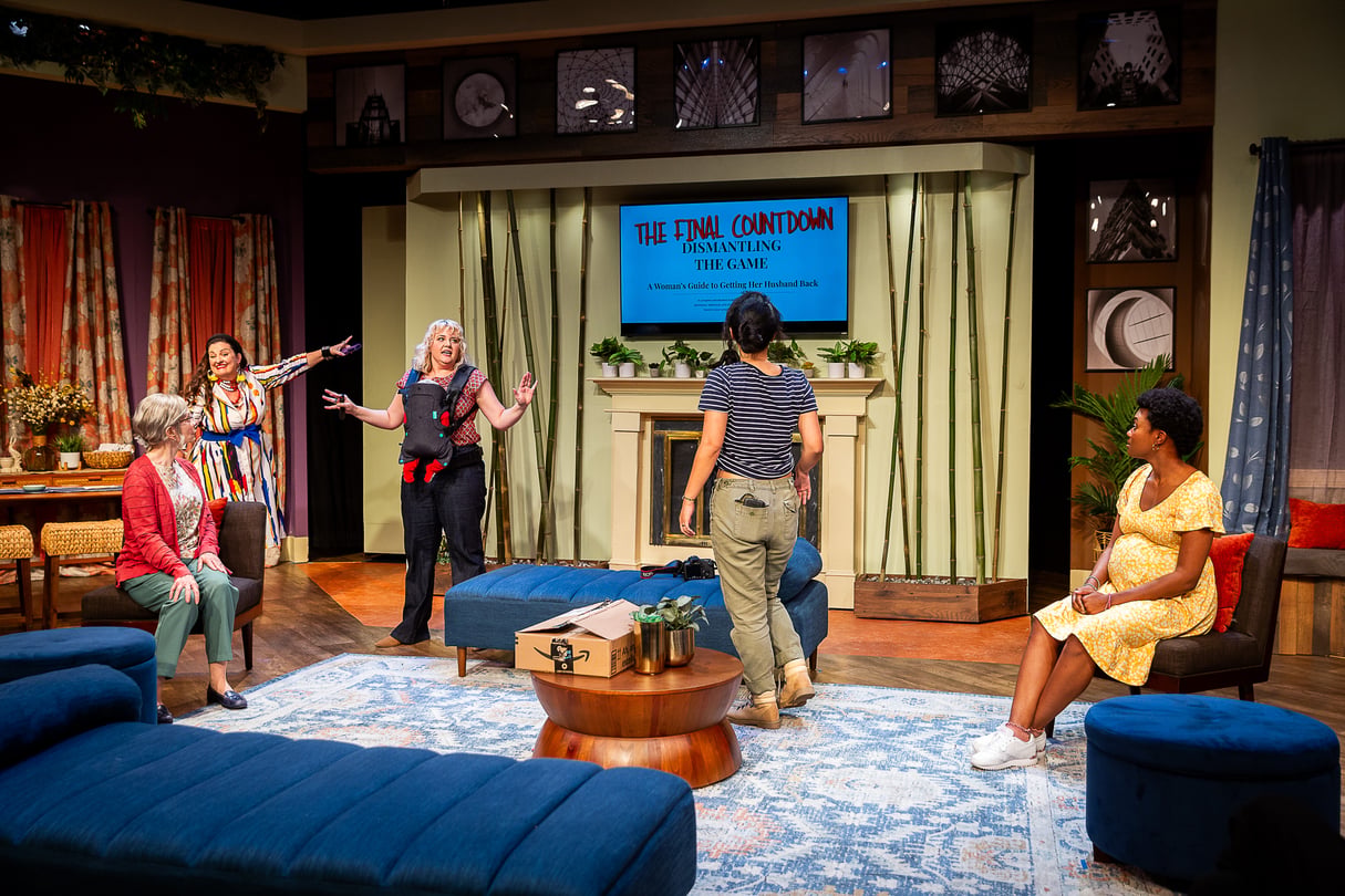 Actors perform on the set of a play in front of a backdrop featuring a fireplace and a TV displaying the words 