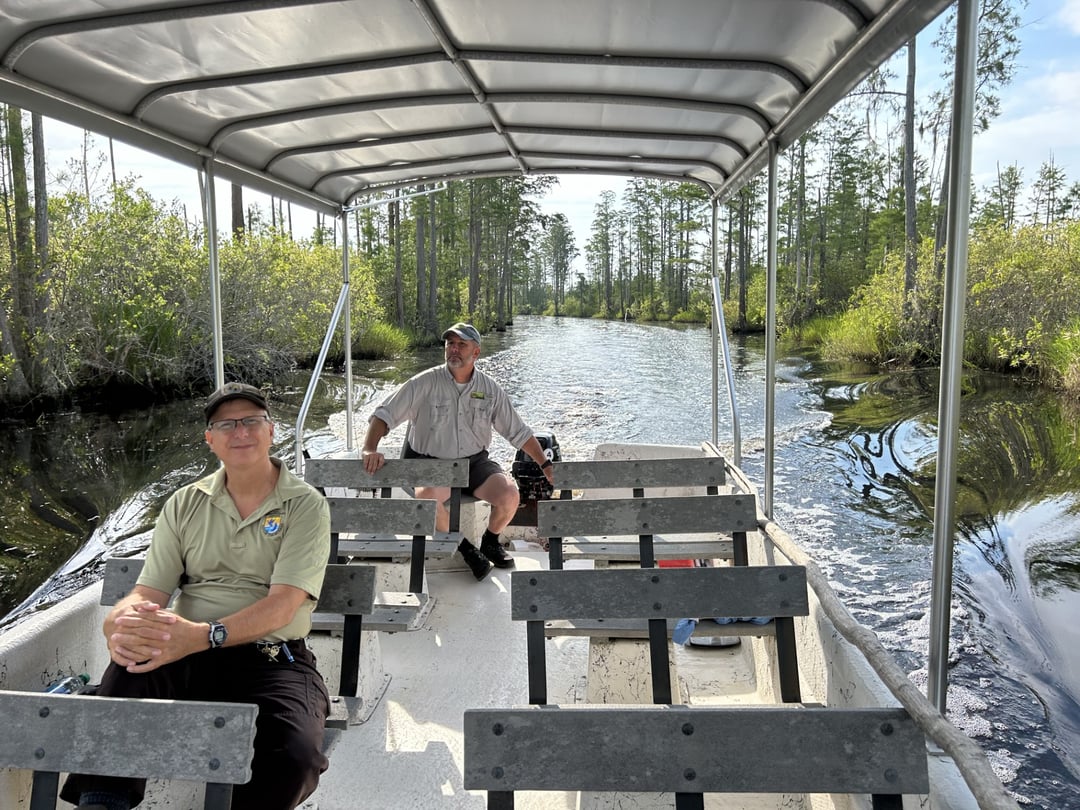 Two men sit on a 24 foot jon boat that has two rows of benches facing the front of the boat. It has a metal cover in top of the boat. In the background is a canal of dark water and green trees on both sides.