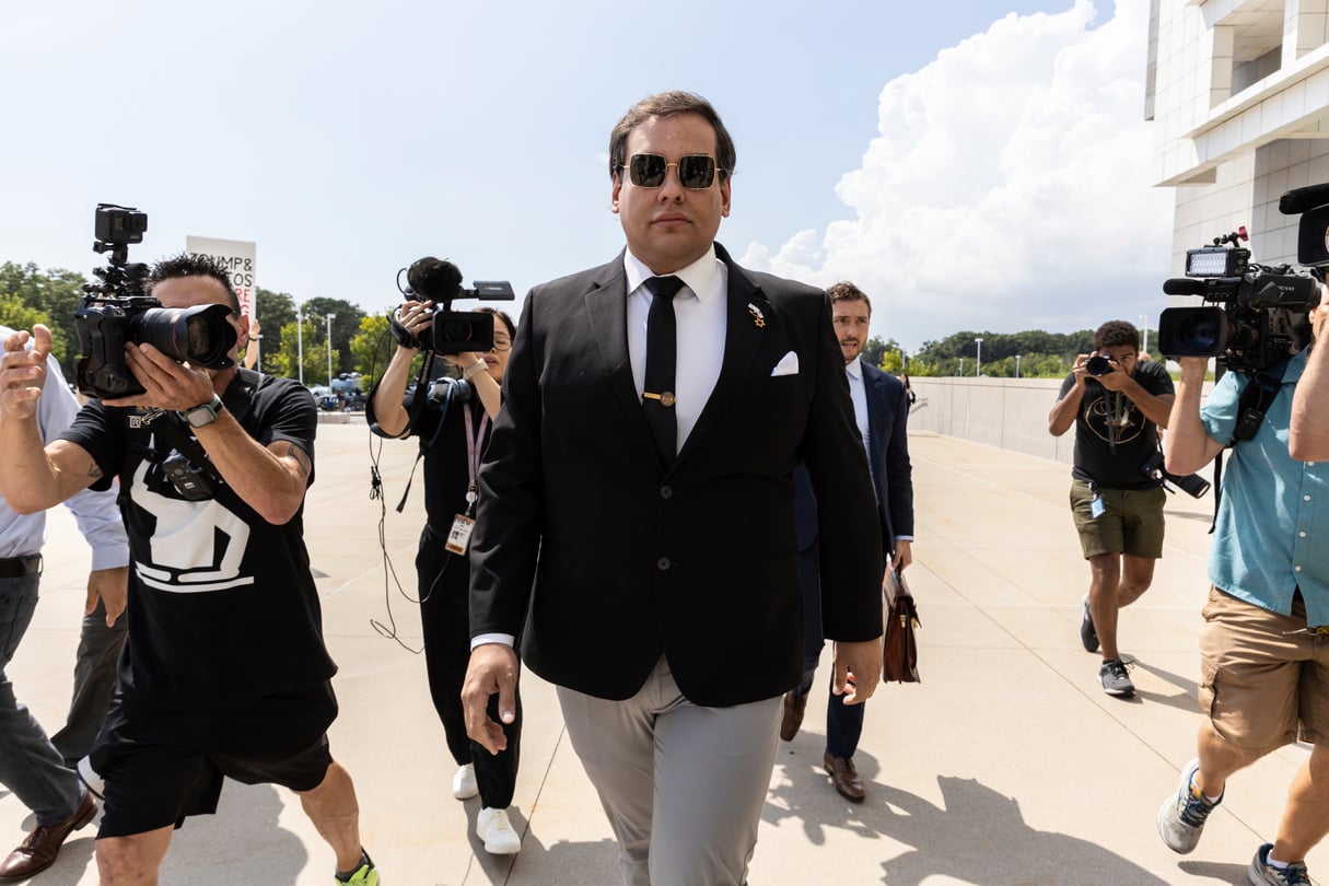Former U.S. Rep. George Santos arrives at court in Central Islip, N.Y. He walks forward, wearing sunglasses, while cameras point at him.