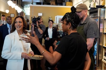 Kamala Harris looks to the side as someone talks to her, and a camera is pointed at her from behind. She is at Dottie's Market in Savannah.