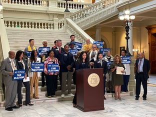 A group of people stand in front of a podium at the bottom of a staircase, holding signs that say We Demand Fair Elections