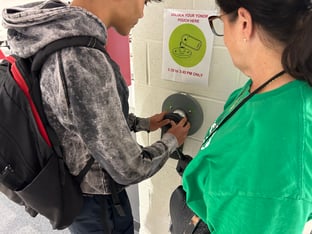 Cheryl Nahmias, wearing a green shirt, stands next to a student unlocking a device pouch.