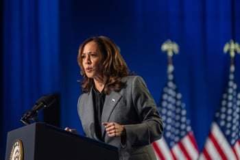 Kamala Harris stands at the podium to deliver remarks at a rally, in front of a deep blue background and several American flags.
