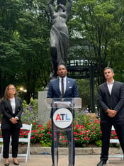 Atlanta Mayor Andre Dickens stands at a podium in front of a statue in Woodruff Park. Cathryn Vassell, left, CEO of Partners for Home, and Frank Fernandez, right, President and CEO at Community Foundation for Greater Atlanta, stand beside him.