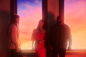 A photo of Khruangbin standing in front of a window with a colorful sunset in the background.